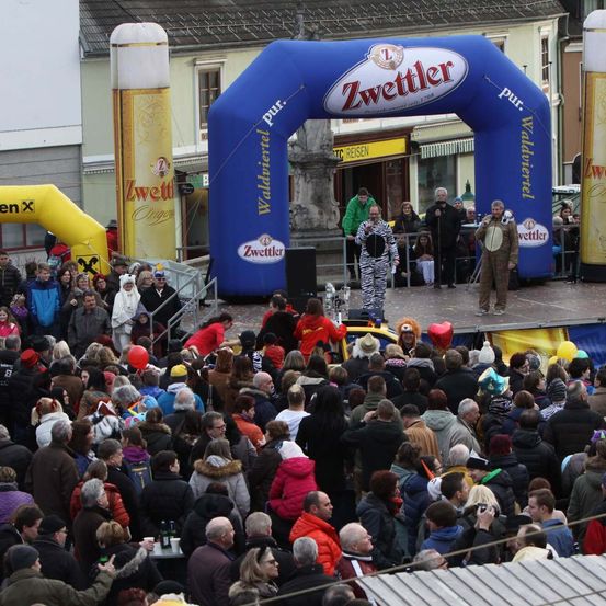 Eine Menschenmenge versammelt sich um eine Bühne, auf der zwei Personen, eine in einem Zebrakostüm und die andere in einer Militäruniform, auftreten. Dahinter befindet sich ein großer blauer Bogen mit der Aufschrift 'Zwettler' und zwei große gelbe aufblasbare Bierkrüge. Im Hintergrund sind Gebäude und ein Steinpfeiler zu sehen.
