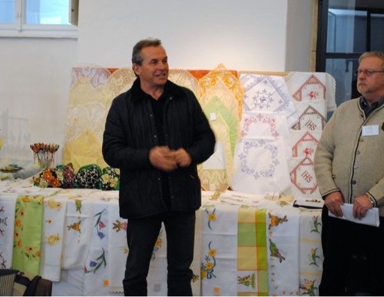 Zwei Männer stehen vor einem Tisch mit gestickten Servietten. Einer gestikuliert. Der Raum hat eine weiße Wand und ein Fenster.