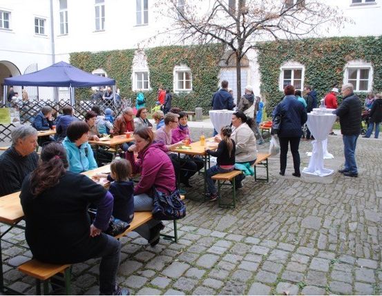 Menschen sitzen an Tischen in einem Innenhof mit Ziegelboden, einem blauen Zelt und einem Gebäude mit weißen Wänden und Efeu.