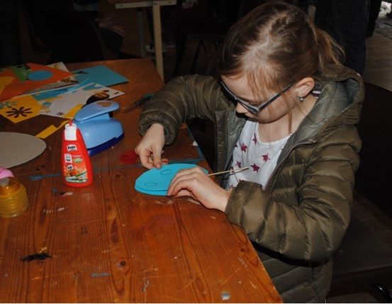 Ein junges Mädchen mit Brille bastelt einen blauen Papierkreis auf einem Holztisch. Verschiedene Bastelmaterialien sind um sie herum verstreut.