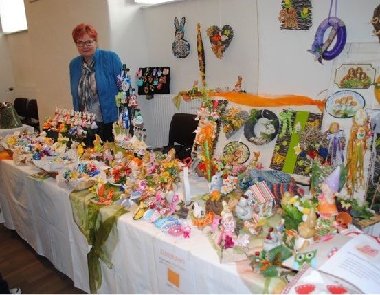 Eine Frau steht hinter einem Tisch, der mit Osterdekorationen bedeckt ist, darunter Figuren, Kerzen und bunte Objekte. Der Raum hat weiße Wände und einen Holzboden.