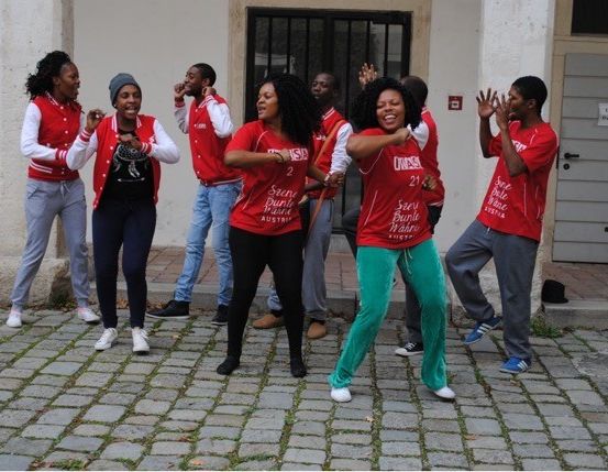 Eine Gruppe von Menschen in roten Shirts tanzt auf einer Kopfsteinpflasterstraße. Sie tragen Turnschuhe und einige tragen Mützen.