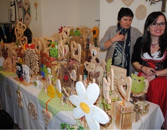 Zwei Frauen stehen neben einem Tisch, der mit handgefertigten Holzdekorationen gefüllt ist, darunter Blumen, Tiere und Herzformen, in einem Raum mit weißer Wand.