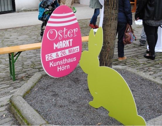 Ein Ostermarktschild steht auf einem Baumstumpf mit einer Hasenausschnitt. Eine Holzbank steht neben dem Schild. Mehrere Leute gehen vorbei.