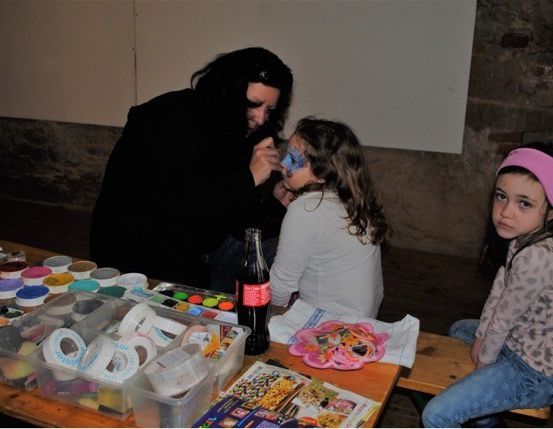 Eine Frau schminkt das Gesicht eines jungen Mädchens, während ein anderes Mädchen in der Nähe sitzt. Sie befinden sich in einem Raum mit einem Holztisch, der mit verschiedenen Kunstmaterialien gefüllt ist.