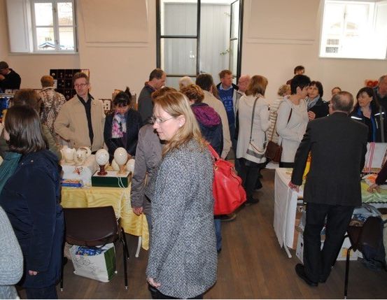 Eine Gruppe von Menschen ist drinnen, vielleicht in einem Laden, und betrachtet Gegenstände auf Tischen. Eine Frau mit Brille schaut auf einen Tisch mit Gegenständen. Ein Mann mit einer Aktentasche steht in der Nähe der Tische.