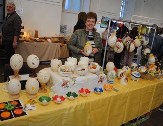 Eine Frau steht vor einem Tisch, auf dem dekorative Eier ausgestellt sind, mit anderen Leuten um sie herum in einem Raum.