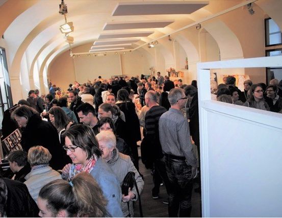 Eine große Menschenmenge, vermutlich bei einer Veranstaltung, versammelt sich in einem Innenraum. Einige Personen unterhalten sich, während andere die Umgebung beobachten. Der Bereich ist gut beleuchtet mit Deckenlampen.