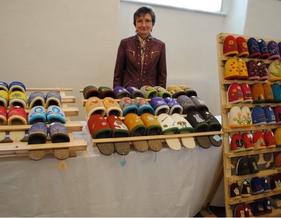Eine Frau steht hinter einem Tisch, auf dem eine Vielzahl bunter Hausschuhe ausgestellt ist. Die Hausschuhe sind auf Holzpaletten und Regalen angeordnet.