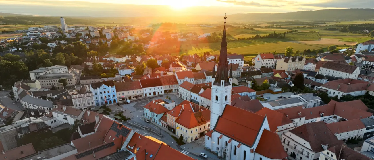 Luftaufnahme einer Stadt mit roten Dächern, einer auffälligen Kirche mit einem Turm und grünen Feldern. Die Sonne geht hinter dem Horizont unter und wirft ein warmes Licht.