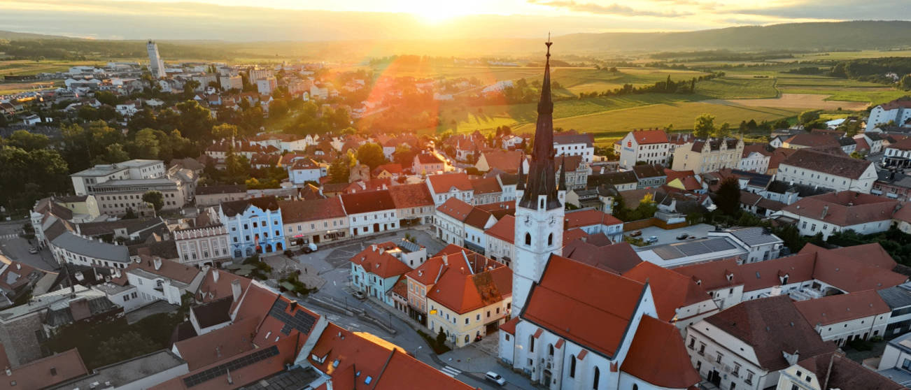 Luftaufnahme einer Stadt mit roten Dächern, einer auffälligen Kirche mit einem Turm und grünen Feldern. Die Sonne geht hinter dem Horizont unter und wirft ein warmes Licht.