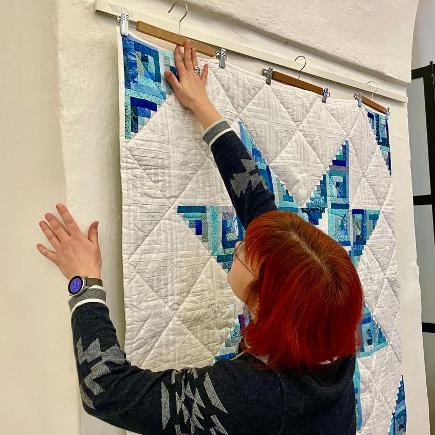 Eine Frau mit roten Haaren hängt eine Decke an einer Wand mit einem Holzaufhänger auf. Die Decke hat ein geometrisches Muster in Blau und Weiß.