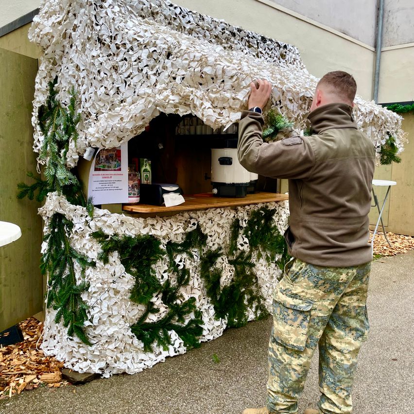 Ein Mann in einer Militäruniform repariert einen getarnten Kiosk mit Dach. Im Inneren des Kiosks befinden sich Flaschen und eine Kaffeemaschine. Der Kiosk ist mit grünem Moos und Tannenzweigen dekoriert.