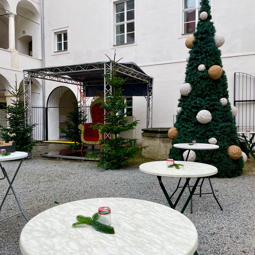 Ein Innenhof mit Weihnachtsbaum, runden Tischen und einem Zeltdach. Ein roter Stuhl steht unter dem Dach.