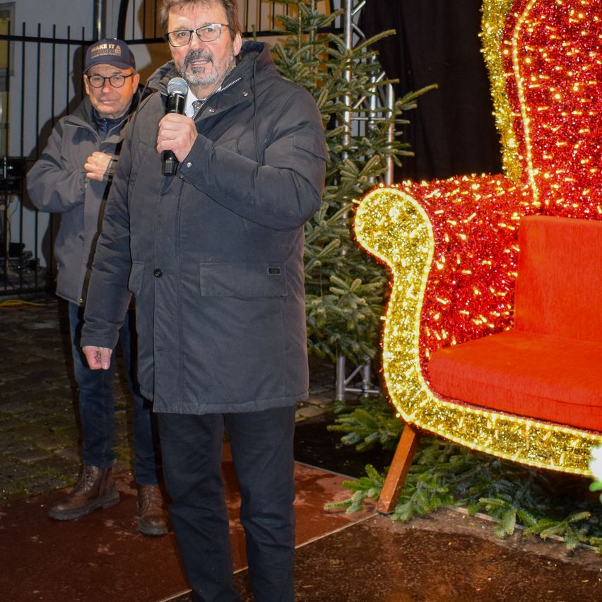 Ein Mann steht neben einem dekorierten Stuhl und spricht in ein Mikrofon. Ein anderer Mann steht hinter ihm und trägt einen Hut und eine Brille. Sie befinden sich in einem Außenbereich mit einem Weihnachtsbaum und einem Metallzaun im Hintergrund.