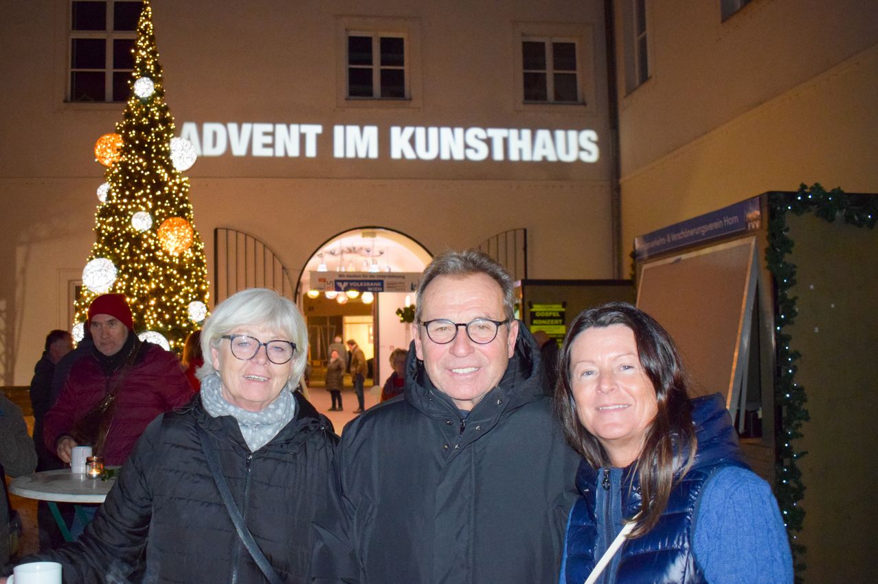 Drei Menschen lächeln vor einem Kunsthaus-Gebäude mit einem beleuchteten Weihnachtsbaum und einem Schild, das Advent anzeigt.