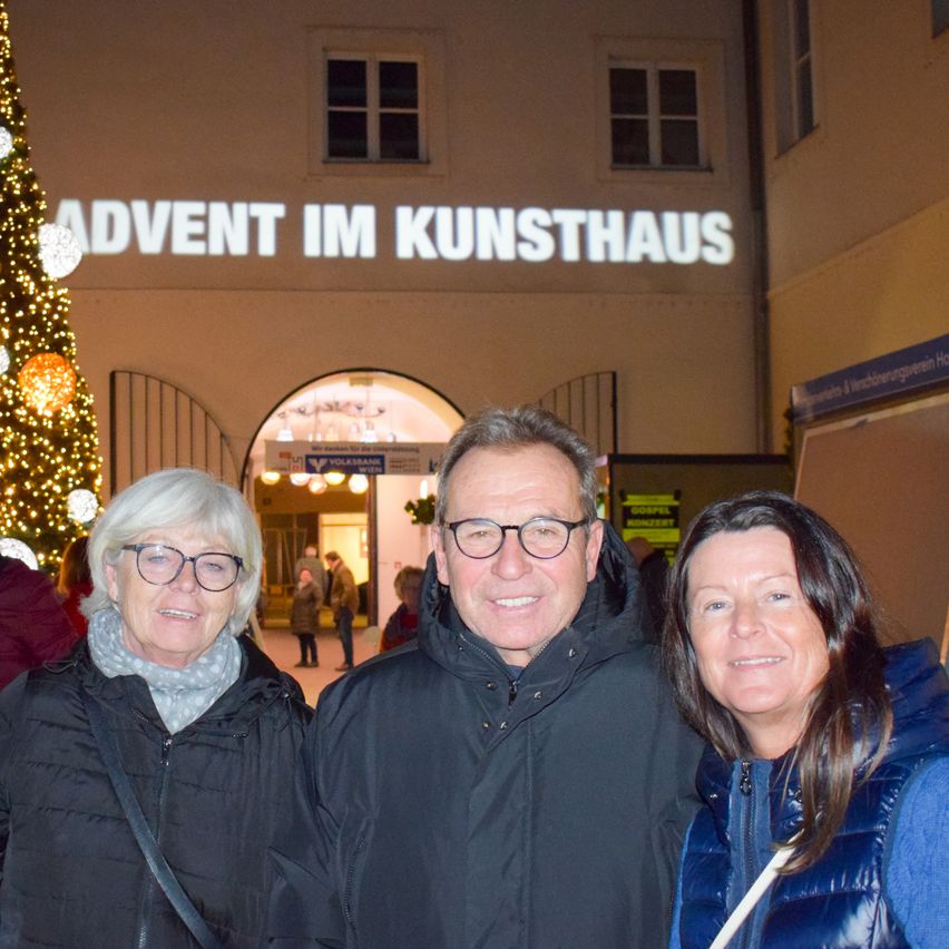 Drei Menschen lächeln vor einem Kunsthaus-Gebäude mit einem beleuchteten Weihnachtsbaum und einem Schild, das Advent anzeigt.