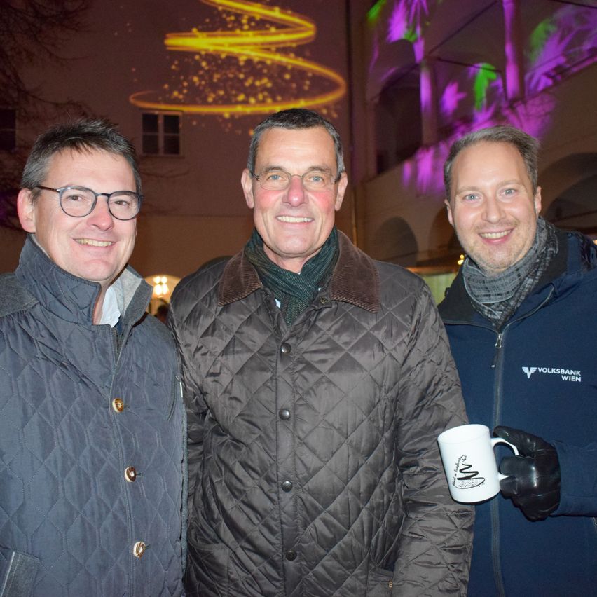 Drei Männer lächeln und posieren für ein Foto vor einem dekorierten Gebäude bei Nacht. Der Mann auf der linken Seite trägt eine Brille und eine Jacke. Der Mann in der Mitte trägt einen Schal und eine Jacke. Der Mann auf der rechten Seite hält eine Tasse.
