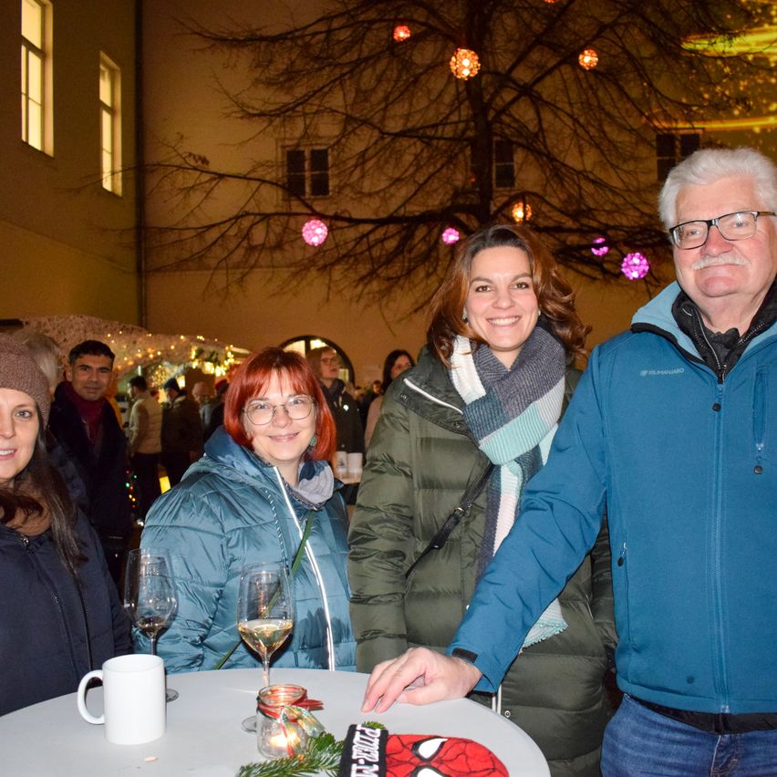 Vier Erwachsene stehen um einen Tisch bei einer Veranstaltung im Freien. Ein Mann und eine Frau tragen eine Brille. Der Tisch hat eine weiße Tasse, Weingläser und eine Kerze. Dahinter ist ein Baum mit Lichtern geschmückt.