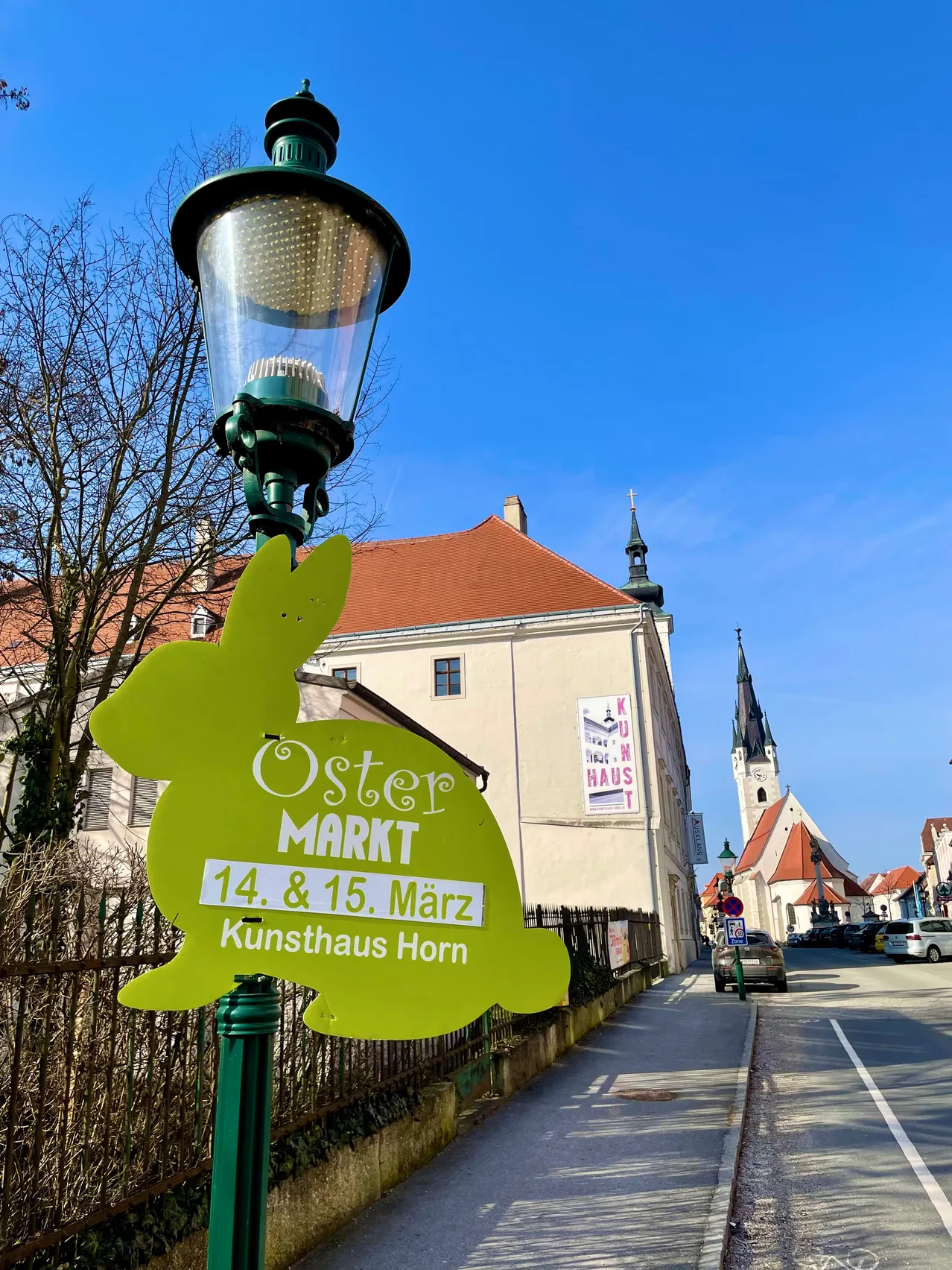 Eine Straßenlaterne zeigt ein grünes, hasenförmiges Schild, das einen Ostermarkt am 14. und 15. März im Kunsthaus Horn ankündigt. Im Hintergrund befinden sich Gebäude mit roten Dächern und einem Turm unter einem klaren blauen Himmel.