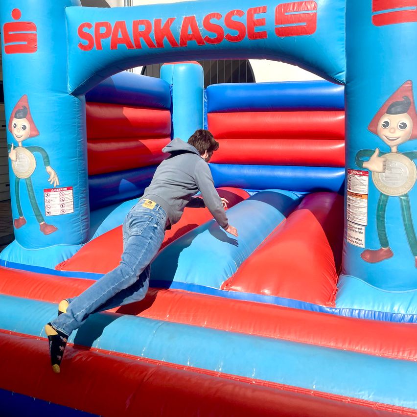 Eine Person spielt in einem Sprungburg mit dem Wort 'Sparkasse' darauf. Die Person trägt eine Kapuzenjacke und Jeans.