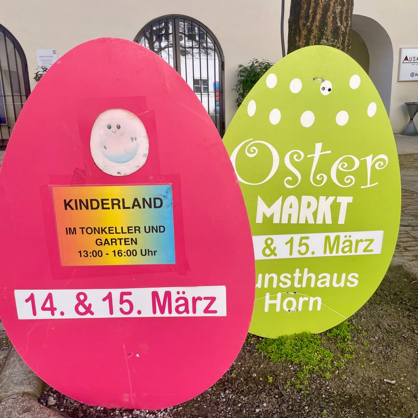 Zwei farbenfrohe eiförmige Schilder vor einem Gebäude. Das rosa Schild sagt 'Kinderland' und hat einen Zeitplan für den 14. und 15. März. Das grüne Schild steht für 'Oster Markt' und erwähnt den 15. März.
