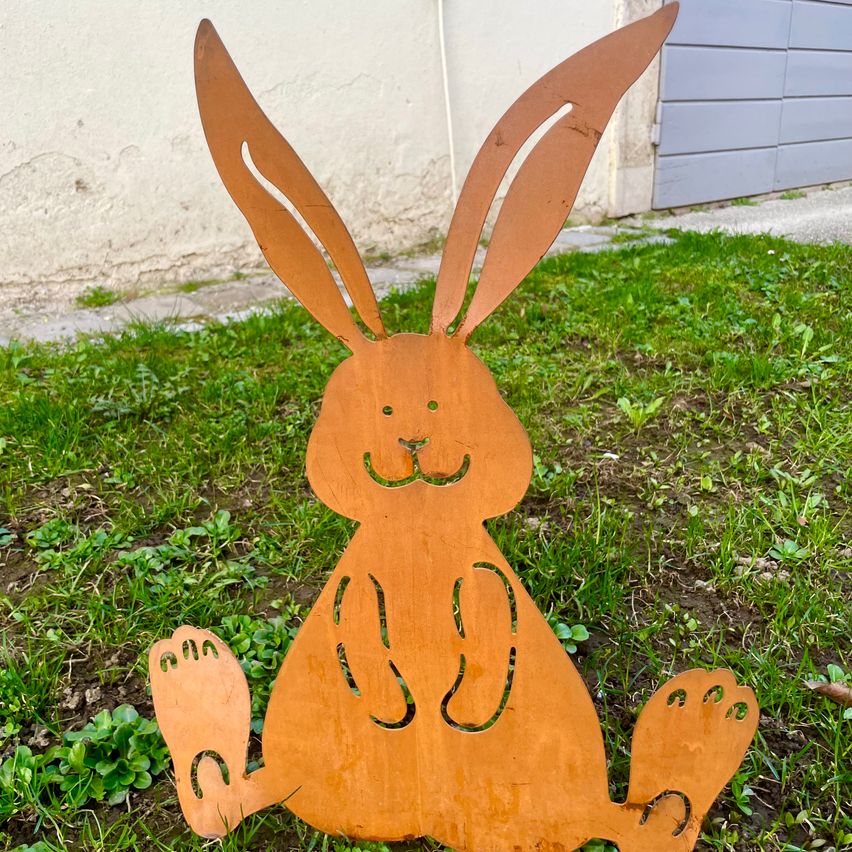 Ein Metallausschnitt eines Kaninchens steht auf einer Rasenfläche. Das Kaninchen hat große Ohren und sitzt auf dem Gras. Der Boden hat etwas Erde und Pflanzen. Hinter dem Kaninchen befindet sich eine Wand und eine Garage.