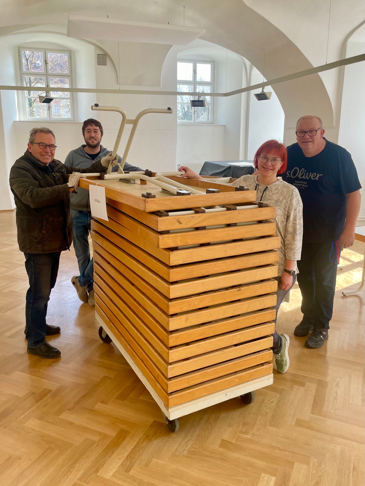 Vier Personen stehen um einen Stapel hölzerner Kisten auf Rädern in einem Raum mit Holzböden und weißen Wänden.