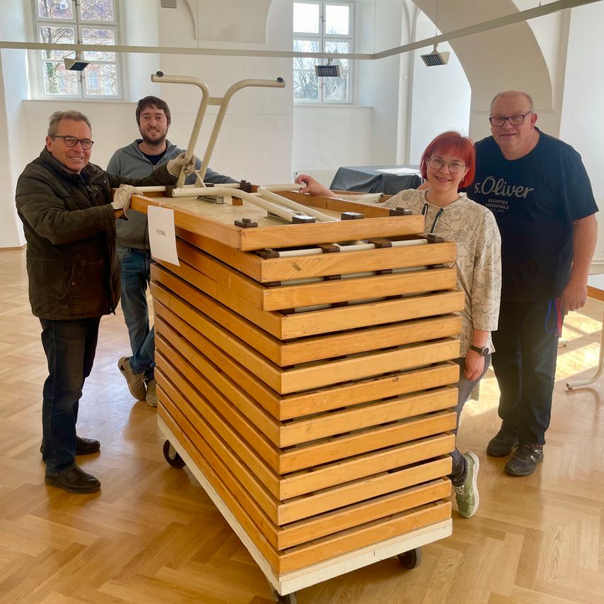 Vier Personen stehen um einen Stapel hölzerner Kisten auf Rädern in einem Raum mit Holzböden und weißen Wänden.