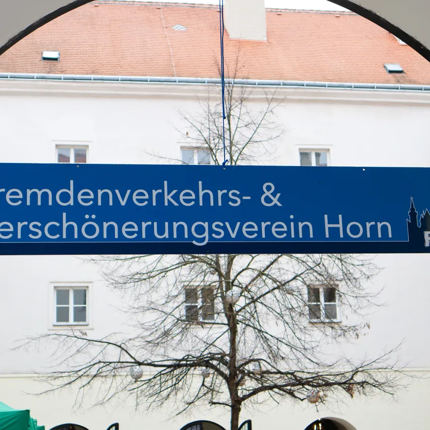 Ein blaues Schild über einem Bogen steht Fremdenverkehrs- & Verschönerungsverein Horn. Ein kahler Baum steht vor einem weißen Gebäude.