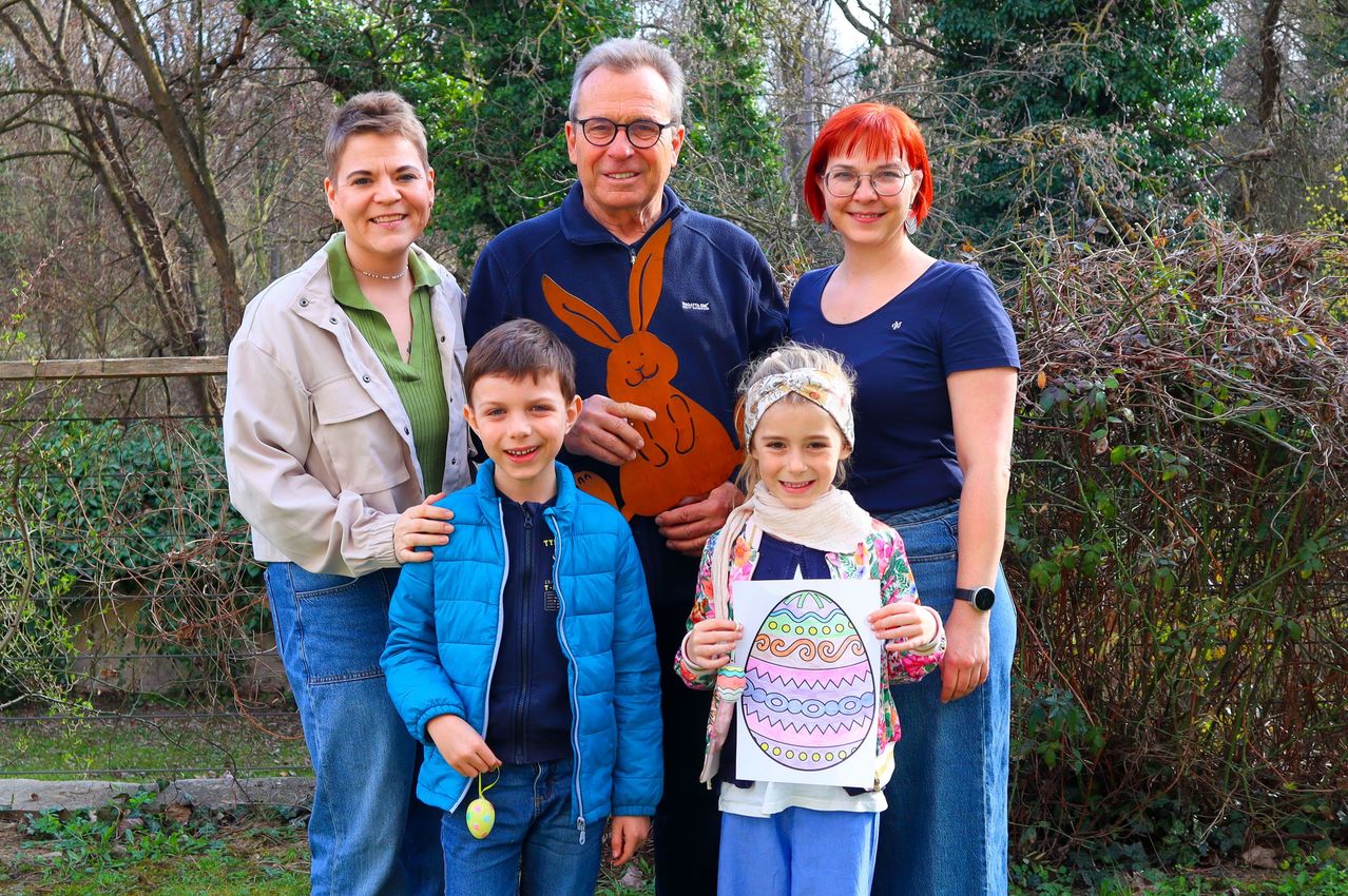 Eine vierköpfige Familie, bestehend aus zwei Erwachsenen und zwei Kindern, steht in einem Garten. Die Erwachsenen tragen Freizeitkleidung, und die Kinder halten bunte Ostereier. Der Mann in der Mitte trägt eine blaue Jacke mit einem Hasendesign.