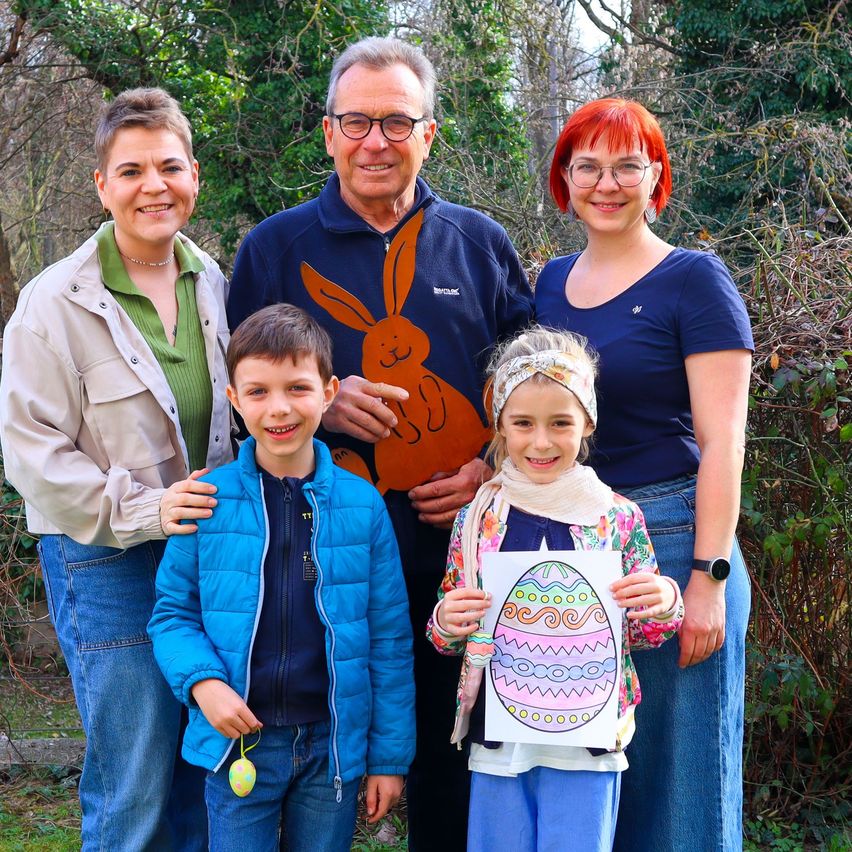 Eine vierköpfige Familie, bestehend aus zwei Erwachsenen und zwei Kindern, steht in einem Garten. Die Erwachsenen tragen Freizeitkleidung, und die Kinder halten bunte Ostereier. Der Mann in der Mitte trägt eine blaue Jacke mit einem Hasendesign.