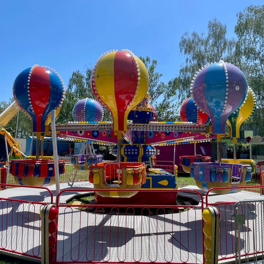 Ein buntes Heißluftballon-Karussell in einem Vergnügungspark. Das Karussell hat mehrere Ballons in Rot, Gelb, Blau und Lila. Es steht auf einer Plattform mit einer roten Barriere.