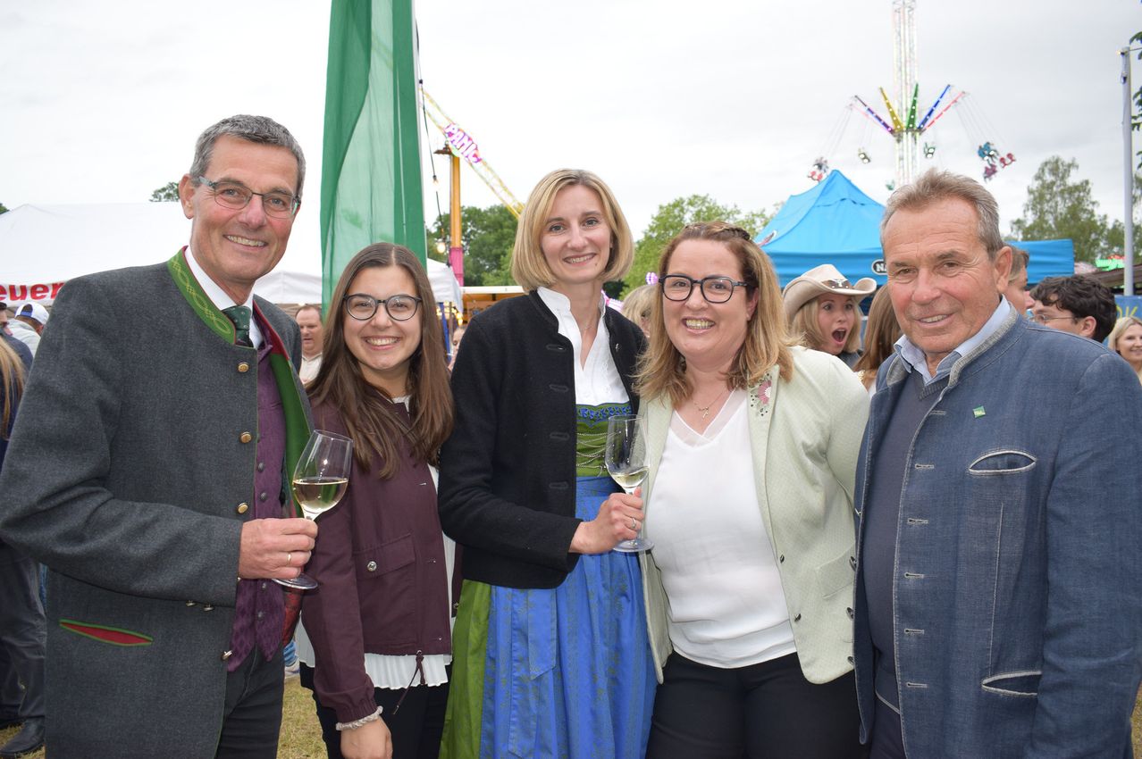Fünf Menschen stehen bei einer Outdoor-Veranstaltung zusammen. Ein Mann und eine Frau halten Weingläser. Eine weitere Frau trägt ein traditionelles bayerisches Kleid. Hinter ihnen sind bunte Fahrgeschäfte und Zelte.