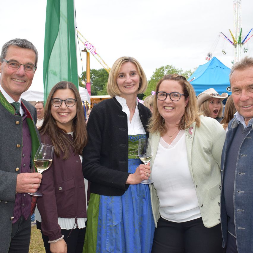 Fünf Menschen stehen bei einer Outdoor-Veranstaltung zusammen. Ein Mann und eine Frau halten Weingläser. Eine weitere Frau trägt ein traditionelles bayerisches Kleid. Hinter ihnen sind bunte Fahrgeschäfte und Zelte.