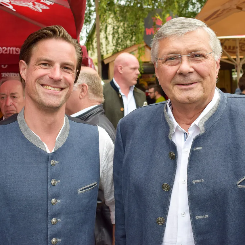 Zwei Männer stehen nebeneinander lächelnd, traditionelle deutsche Kleidung mit dekorativen Knöpfen tragen. Sie sind in einem Außenbereich mit Leuten und Zelten im Hintergrund.