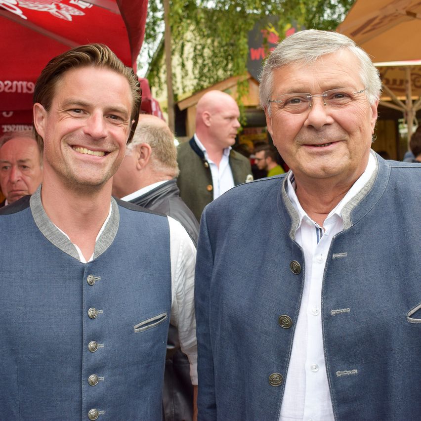 Zwei Männer stehen nebeneinander lächelnd, traditionelle deutsche Kleidung mit dekorativen Knöpfen tragen. Sie sind in einem Außenbereich mit Leuten und Zelten im Hintergrund.