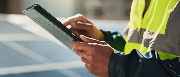 Eine Person in einer Sicherheitsweste hält ein Tablet vor Solarmodulen und überprüft oder überwacht sie wahrscheinlich.