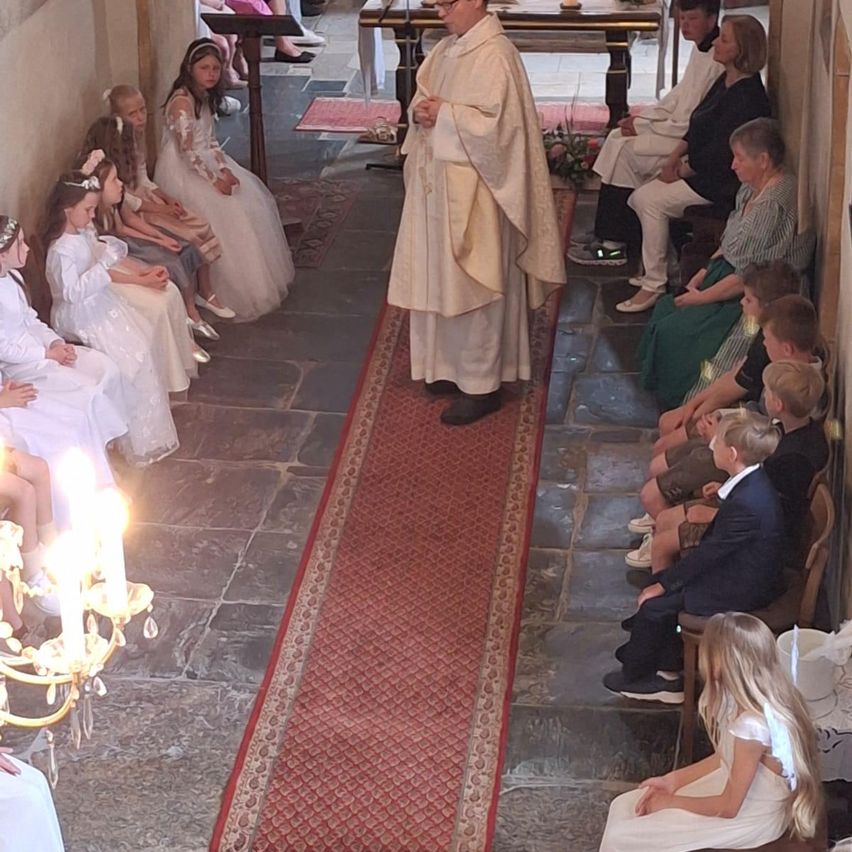 Ein Priester steht in einer Kirche und blickt auf eine Gruppe von Kindern in weißen Kleidern, die auf Steinstufen sitzen. Sie sitzen mit Erwachsenen, und ein roter Teppich führt zum Altar.