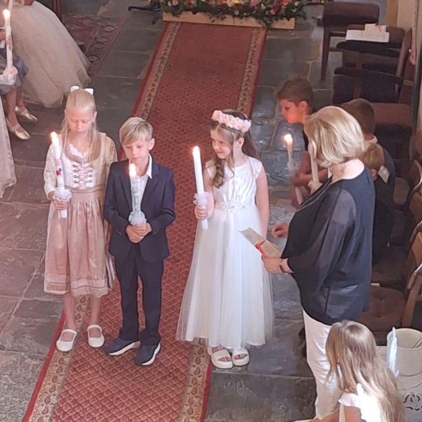 Eine Gruppe von Kindern, möglicherweise bei einer Hochzeit, hält brennende Kerzen. Eine Frau hält ein Büchlein. Dahinter sitzen ein Bräutigam und eine Braut.