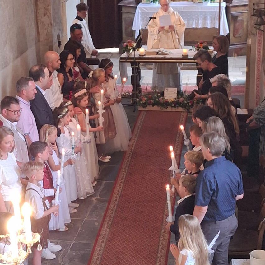Eine Gruppe von Menschen versammelt sich in einer Kirche und hält Kerzen. Ein Priester steht am Altar. Kinder und Erwachsene, in Weiß gekleidet, stehen in Reihen. Die Kirche ist mit Blumen und Kerzen geschmückt.