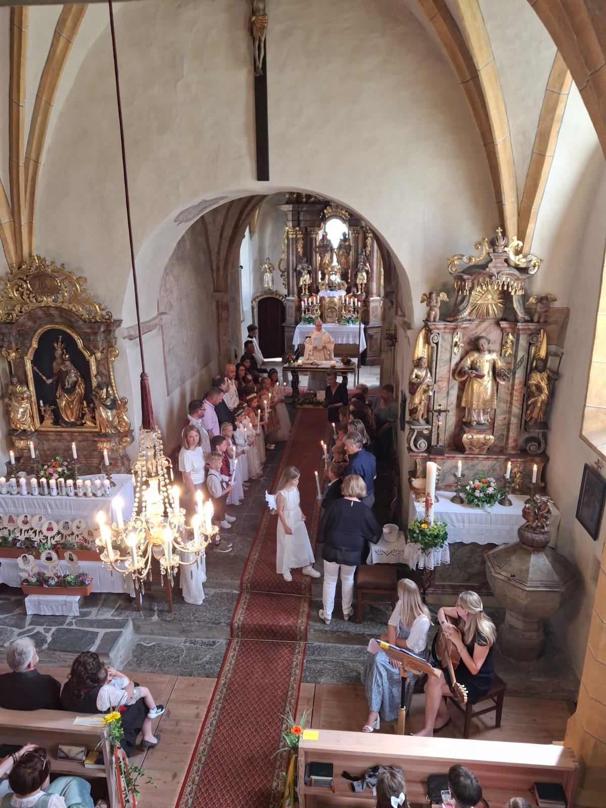 Ein Kircheninneres mit einem großen Bogen, kunstvollen Altar und einer Gruppe von Menschen mit Kerzen. Ein Kronleuchter und Blumen schmücken den Altar, während Statuen an den Wänden zu sehen sind.