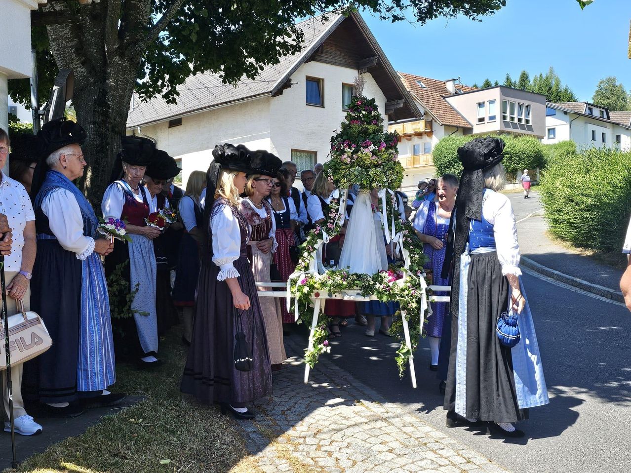 Eine Gruppe von Frauen in traditionellen Kostümen steht um einen geschmückten Wagen in einem Dorf. Eine Frau trägt ein weißes Hochzeitskleid auf dem Wagen.