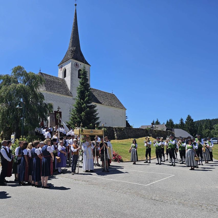 Eine Gruppe von Menschen in traditioneller Kleidung steht vor einer Kirche an einem sonnigen Tag. Ein Priester führt eine Prozession an.