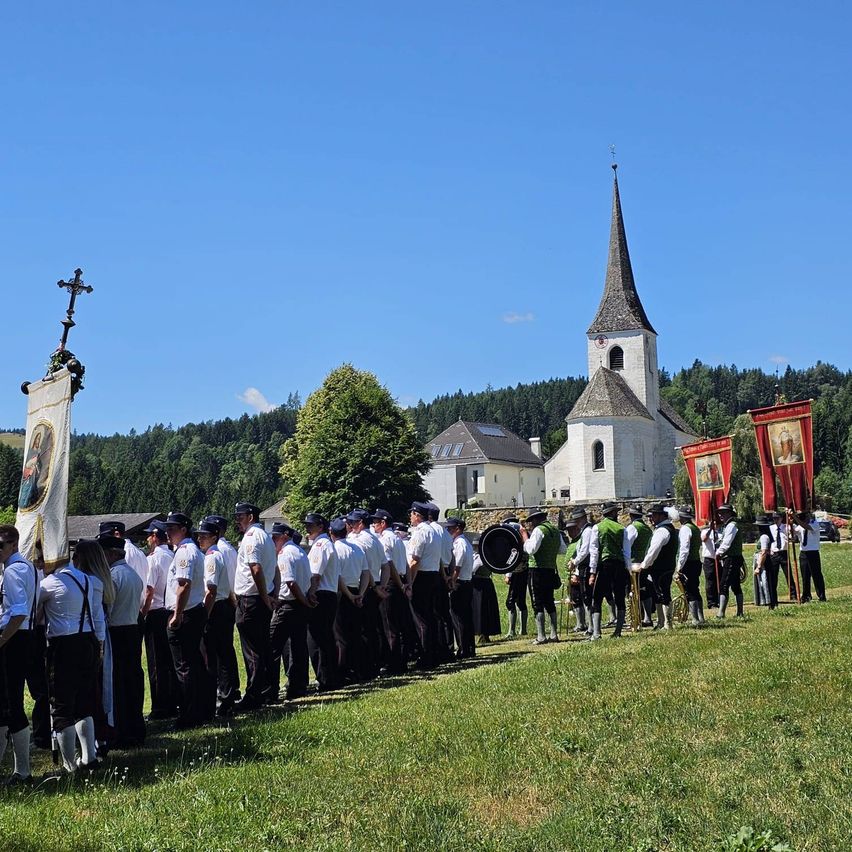 Eine Gruppe von Menschen in traditioneller Kleidung steht in einer Reihe vor einer Kirche mit einem Turm unter einem klaren blauen Himmel.