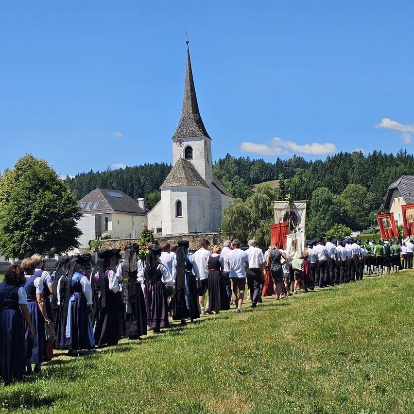 Eine Gruppe von Menschen in traditioneller Kleidung steht in einer Reihe vor einer Kirche mit Turmspitze unter einem klaren blauen Himmel.