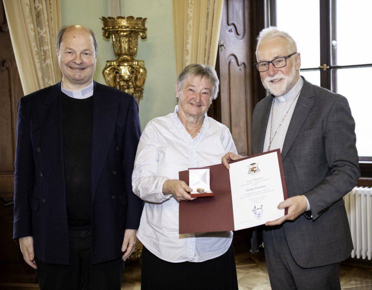 Drei Personen posieren für ein Foto. Eine Frau hält ein Zertifikat und eine Medaille. Zwei Männer stehen neben ihr und lächeln.