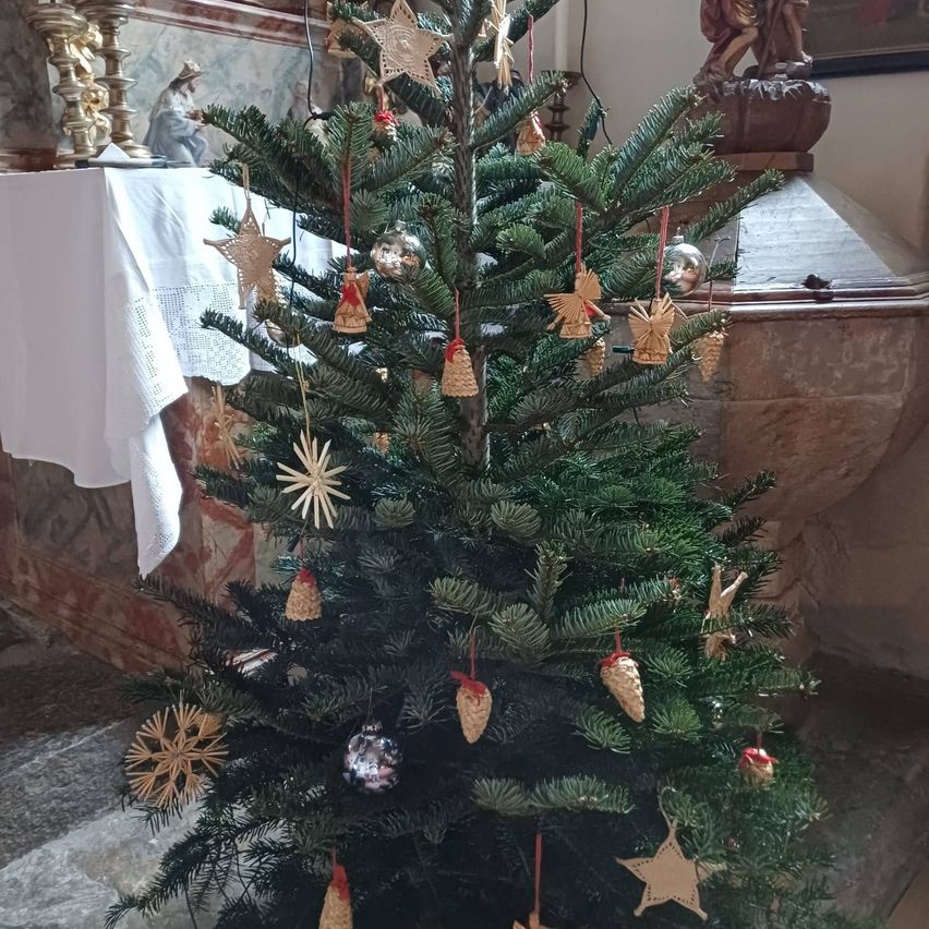 Ein Weihnachtsbaum mit verschiedenen Ornamenten wie Sternen, Engeln und Tannenzapfen. Er steht in einem Raum mit Steinwänden und einem mit einem weißen Tuch bedeckten Tisch.