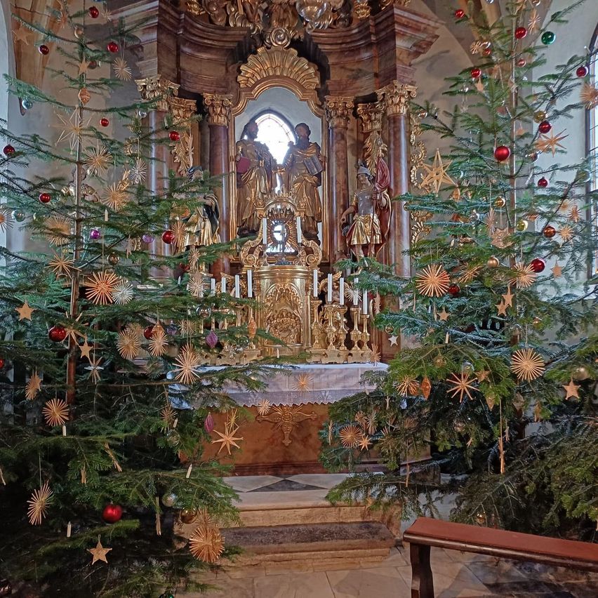 Zwei große Weihnachtsbäume, geschmückt mit Ornamenten und Sternen, flankieren den prächtigen Altar einer Kirche. Der Altar ist golden und hat einen Spiegel darüber mit Statuen.