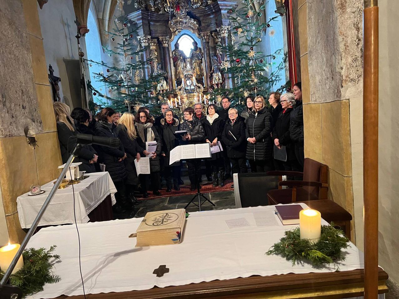 Ein Chor steht vor einem Kirchenaltar, mit Weihnachtsbäumen im Hintergrund. Der Chor hält Notenblätter in der Hand und steht vor einer Kerze und einem Buch auf einem Tisch.